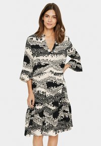 Mustat ja valkoiset kukkakuosiset mekko, jossa on V-pääntie, laineilevat hihat ja kerroksellinen helma. Valmistettu kevyestä kankaasta.