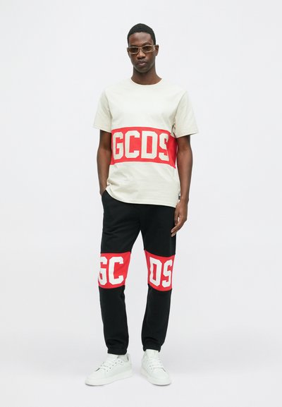 T-shirt in cotone beige con grafica "GCDS" rossa e bianca, abbinata a joggers neri con dettagli rossi e bianchi e sneakers bianche.