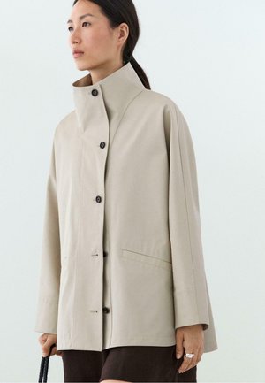 Femme portant un manteau beige boutonné à col montant, tenant un sac à poignée tressée noire, sur un fond clair uni.
