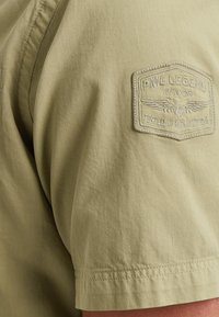 Khaki short-sleeve shirt made of textured fabric featuring an embroidered patch on the left shoulder with "PME Legend Aviator" and wing design.