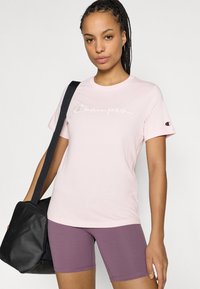 Ροζ κοντομάνικη μπλούζα με το λογότυπο "Champion" κεντημένο σε γραφ ή. Συνδυάζεται με μωβ σορτς και μαύρη τσάντα ώμου.