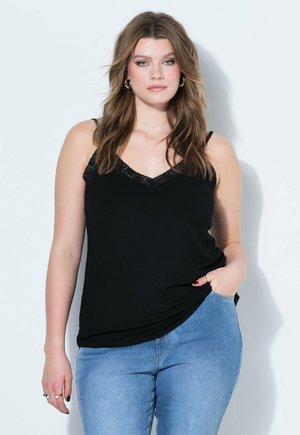 Junge Frau mit welligem braunem Haar, die ein schwarzes, ärmelloses Oberteil mit Spitzenbesatz und hellblaue Jeans trägt, vor einem weißen Hintergrund stehend.