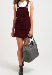 Bordowa sukienka ogrodniczka z materiału sztruksowego z regulowanymi ramiączkami, zestawiona z szarym skórzanym torebką oraz czarnymi botkami za kostkę.