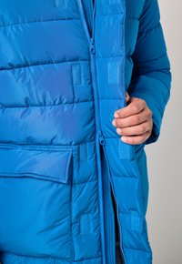 Giacca a vento blu con motivo trapuntato, chiusura con zip e dettagli in velcro. Presenta pratiche tasche laterali e una texture liscia.