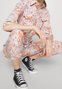 Jumper floral en tonos pastel con acentos en naranja y verde, que presenta mangas cortas, parte delantera con botones y puños elásticos. Combinado con zapatillas negras de Converse.