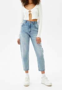 Nainen, jolla on päällään valkoinen lyhyt pitkähihaisena paita, jossa on leikkaus, ja korkeavyötäröiset vaaleansiniset farkut, valkoiset sukat ja valkoiset platofom-tennarit.