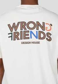 Camiseta blanca con el texto "WRONG FRIENDS" en varios colores y tipografías, con "DESIGN HOUSE" abajo. La tela parece suave.
