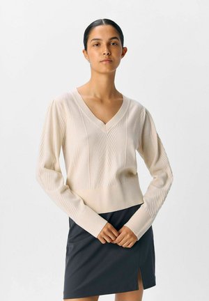 Hellbeige gestrickter Pullover mit V-Ausschnitt, strukturiertem Muster und gerippten Bündchen, kombiniert mit einem dunklen Rock, der eine taillierte Silhouette betont.