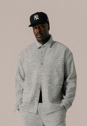 Grau strukturierte Jacke mit Knopfverschluss, seitlichen Taschen und lässigem Schnitt, kombiniert mit passenden Hosen und einer schwarzen Mütze.