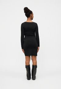 Μαύρο φόρεμα με μακριά μανίκια σε εφαρμοστή γραμμή, που φτάνει μέχρι το μέσο του μηρού. Φοράει ψηλές μαύρες μπότες, εμφανιζόμενο από πίσω πάνω σε απλό φόντο.