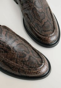 Ruskeat käärmeennahkakuosiset loaferit, joissa on sileä pinta, pyöreä kärki ja kontrastivärinen musta pohja; teksturoitu kuosi lisää syvyyttä ja visuaalista kiinnostavuutta.
