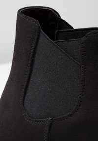 Bottine noire avec un dessus en daim doux et des panneaux latéraux élastiques, présentant une texture lisse et des détails de couture minimaux.