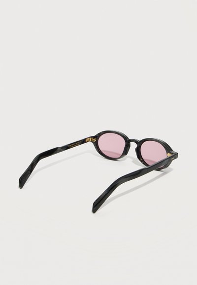 Μαύρα οβάλ γυαλιά ηλίου με λεπτά πλαίσια και ανοιχτό ροζ φακούς, ανοιχτά πάνω σε λευκή επιφάνεια.