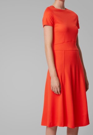 Femme portant une robe ajustée orange vif à manches courtes et longueur genoux, avec une jupe évasée sur fond gris uni.