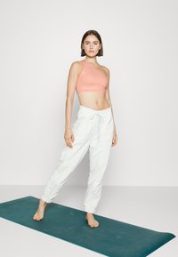 Top cropped rosa de alça única, combinado com calças de jogging brancas soltas. Modelo descalço em um tapete de yoga teal. Estilo simples e casual de athleisure.