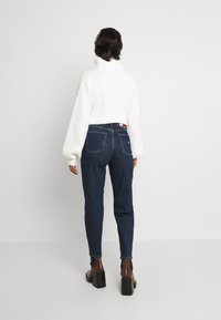 Vit stickad tröja med voluminösa ärmar, i kombination med mörkblå jeans. Modellen bär bruna klackskor med blockklack.