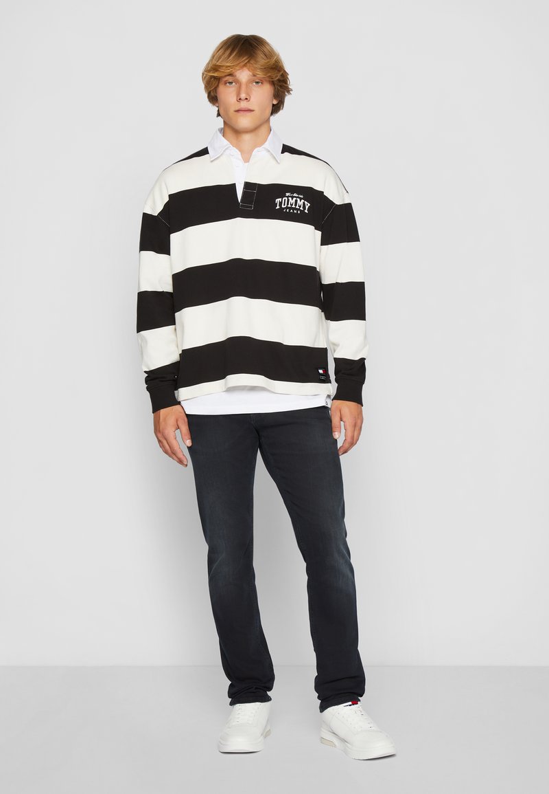 Czarno-biały pasiasty rugby z długim rękawem, z kołnierzykiem i logo, noszony na białej koszuli, w zestawieniu z ciemnymi dżinsami i białymi trampkami.