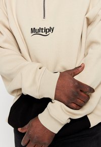 Sweat-shirt beige à fermeture éclair avec logo "Multiply" noir. Présente des poignets côtelés et un ourlet contrastant noir. Tissu doux avec texture visible.