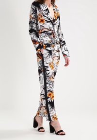 Costume imprimé floral en noir, blanc et orange, comprenant une veste à col cranté et un pantalon ajusté avec des accents sur les côtés. Sandales à talons noires.
