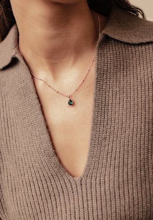 Roségoldene Halskette mit einem tropfenförmigen grünen Anhänger, präsentiert vor einem taubfarbenen Strickpullover mit tiefem V-Ausschnitt.