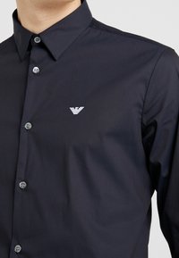 Man med en mörk marinblå skjorta med knappar och spetskrage samt en liten vit broderad örnlogotyp på bröstet.