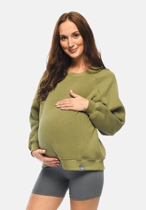 Schwangere Frau mit langen braunen Haaren, die ein olivgrünes Sweatshirt und graue Shorts trägt, hält sanft ihren Bauch und lächelt.