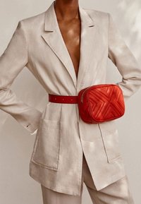 Blazer en lin beige avec un décolleté en V profond, associé à un sac ceinture matelassé rouge. Le sac présente un motif géométrique et un design élégant.
