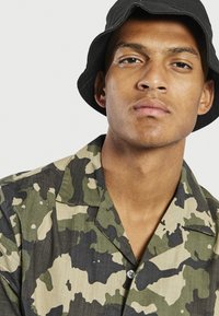 Camicia a maniche corte con bottoni, in un motivo camouflage con tonalità di verde e beige; il tessuto appare leggero e testurizzato. Cappello a secchiello nero.
