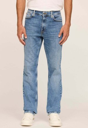 Homme portant un jean droit bleu clair, des baskets blanches et une chemise blanche, se tenant devant un fond beige uni.