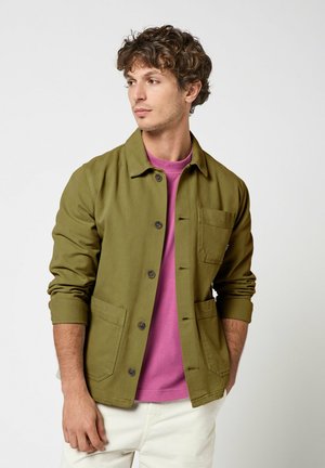 Homme aux cheveux bouclés portant une veste boutonnée vert olive, une chemise rose et un pantalon blanc, regardant vers sa gauche sur un fond uni.