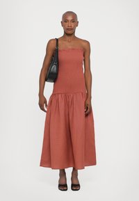 Maxime dress in rust-red with a smocked, strapless bodice and tiered skirt. Smooth texture, paired with a black shoulder bag and open-toe heels.