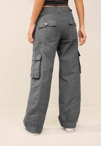Pantalon cargo gris à coupe ample, doté de multiples poches, de boutons en accent et d'un tissu texturé. Le pantalon se resserre aux chevilles.