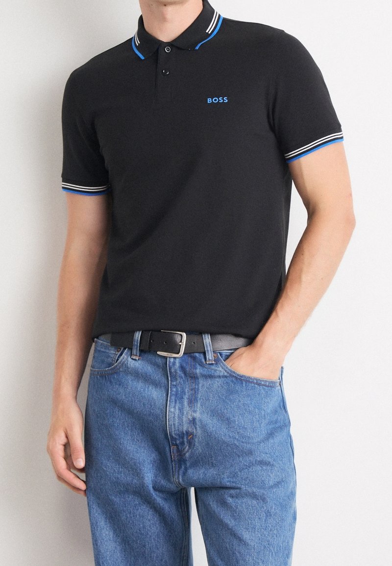 Homme portant un polo noir avec un liseré bleu et blanc et le logo « BOSS », associé à un jean bleu et une ceinture noire, main dans la poche.