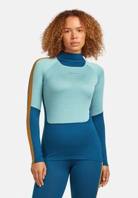 Donna con capelli ricci che indossa un top atletico a maniche lunghe aderente, a blocchi di colore, e leggings abbinati in tonalità di blu e verde acqua chiaro.