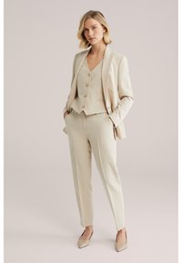 Vrouw staand met handen in de zakken, gekleed in een beige op maat gemaakte blazer, een dichtgeknoopte gilet, bijpassende broek en beige pumps met puntige neus, tegen een lichte achtergrond.