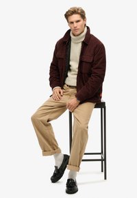 Bordeaux corduroy jasje over een beige coltrui, gecombineerd met licht khaki broeken en zwarte instappers, zittend op een zwarte kruk.