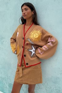 Brun oversized cardigan med røde kanter, med broderier av sol og hjerte, knappeåpning og lange ermer. Kombinert med et beige skjørt.