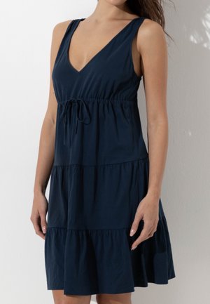 Donna che indossa un vestito senza maniche blu navy con scollo a V e cintura arricciata in vita, in piedi davanti a uno sfondo chiaro.