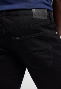 Gros plan d'une personne portant un jean noir Lee et une chemise bleue, montrant la poche arrière et les étiquettes de la marque Lee sur la ceinture et la poche.