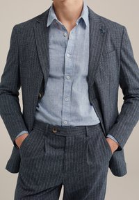 Traje de rayas marino con un ajuste a medida; tejido texturizado; combinado con una camisa de lino azul claro; cuenta con botones y un pañuelo de bolsillo.