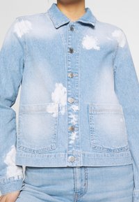 Světle modrá džínová bunda se zapínáním na knoflíky vpředu, dvěma předními kapsami a obláčkovými bílými vzory na látce.