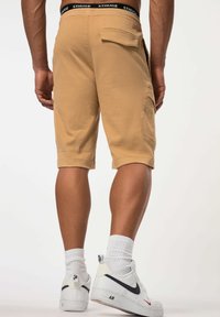 Shorts cargo beiges avec une poche arrière, ourlet roulé et une ceinture noire avec le logo "STHUGE", portés avec des baskets blanches Nike et des chaussettes blanches.