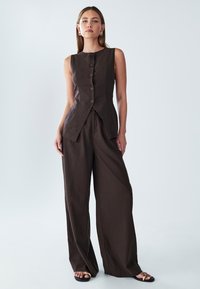 Hnědý top bez ramínek s knoflíky vpředu v kombinaci s širokými hnědými kalhotami. Hladká textura, přizpůsobený střih a minimalistický design.