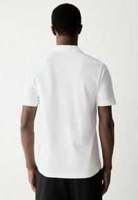 Weißes Kurzarm-Poloshirt mit klassischem Kragen, glatter Baumwollstruktur und seitlichen Schlitzen am Saum. Einfaches, minimalistisches Design.