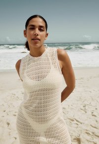 Vestido blanco de ganchillo con diseño sin mangas, que presenta una textura de tejido abierto y un escote con forma de concha, situado frente a un fondo de playa.