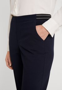 Marinblå byxor i ett slätt tyg, med en sidoficka och midjeband med svart och vit randig detalj.