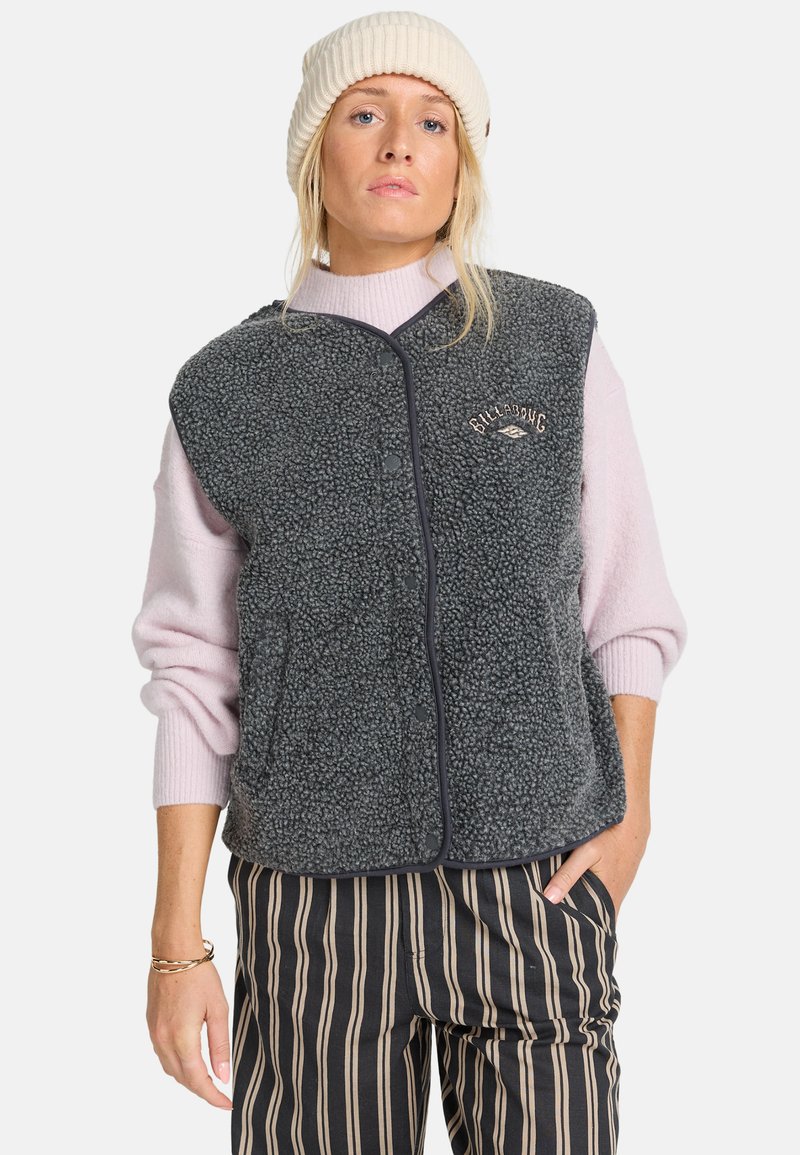 Grijze fleece bodywarmer met een gestructureerde afwerking, drukknoop-front, zwarte accenten en logodetail, gedragen over een lichtroze trui en gestreepte broek.