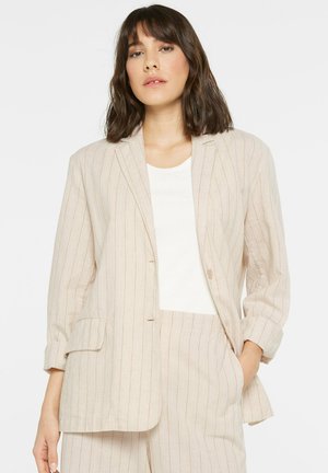 Frau trägt einen beige Nadelstreifen-Blazer und passende Hose mit weißem Oberteil, steht mit einer Hand in der Tasche vor weißem Hintergrund.