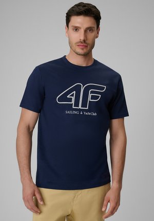 Mężczyzna w granatowej koszuli z białym logo w stylu liny ""4F"" i napisem ""SAILING Yacht Club"", zestawiony z beżowymi spodniami.