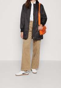 Černá nylonová bombarďácká bunda s žebrovanými manžetami, béžové kordové kalhoty s širokými nohavicemi a bílé tenisky s černými akcenty. Oranžová crossbody taška.